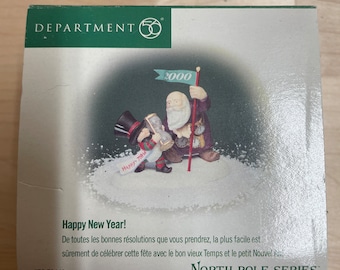 May include: A Department 56 North Pole Series Christmas ornament featuring Santa Claus and a snowman holding a champagne bottle. The ornament is in a box with the text "Happy New Year!" and "2000".
