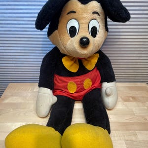K&ouml;nnte beinhalten: Eine Mickey Mouse-Pl&uuml;schpuppe mit schwarzem K&ouml;rper, gro&szlig;en gelben Schuhen und wei&szlig;en Handschuhen. Die Puppe hat eine rote Hose mit gelben Kn&ouml;pfen, eine gelbe Fliege und gro&szlig;e, ausdrucksstarke Augen. Die Puppe sitzt auf einer Holzoberfl&auml;che.