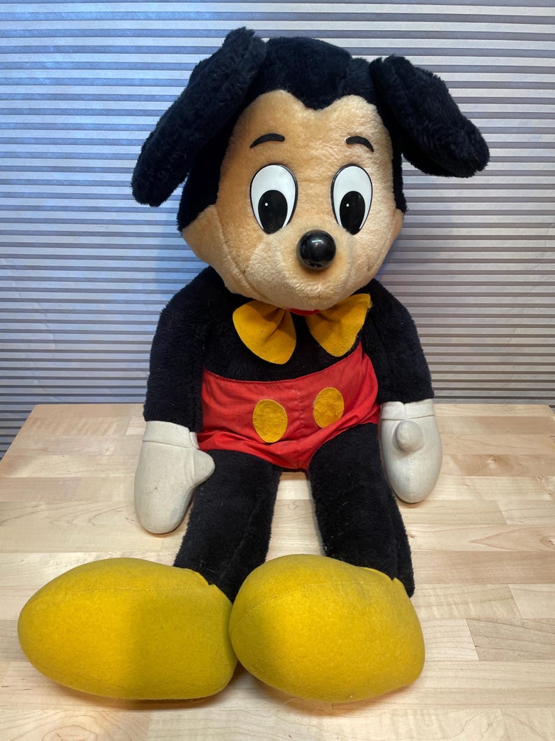 K&ouml;nnte beinhalten: Eine Mickey Mouse-Pl&uuml;schpuppe mit schwarzem K&ouml;rper, gro&szlig;en gelben Schuhen und wei&szlig;en Handschuhen. Die Puppe hat eine rote Hose mit gelben Kn&ouml;pfen, eine gelbe Fliege und gro&szlig;e, ausdrucksstarke Augen. Die Puppe sitzt auf einer Holzoberfl&auml;che.