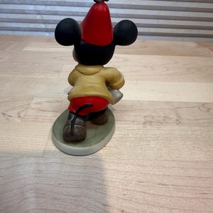 May include: A ceramic figurine of Mickey Mouse from the back, wearing a red hat, yellow jacket, and red shorts. The figure is standing on a green oval base. The figurine has black ears and brown shoes.