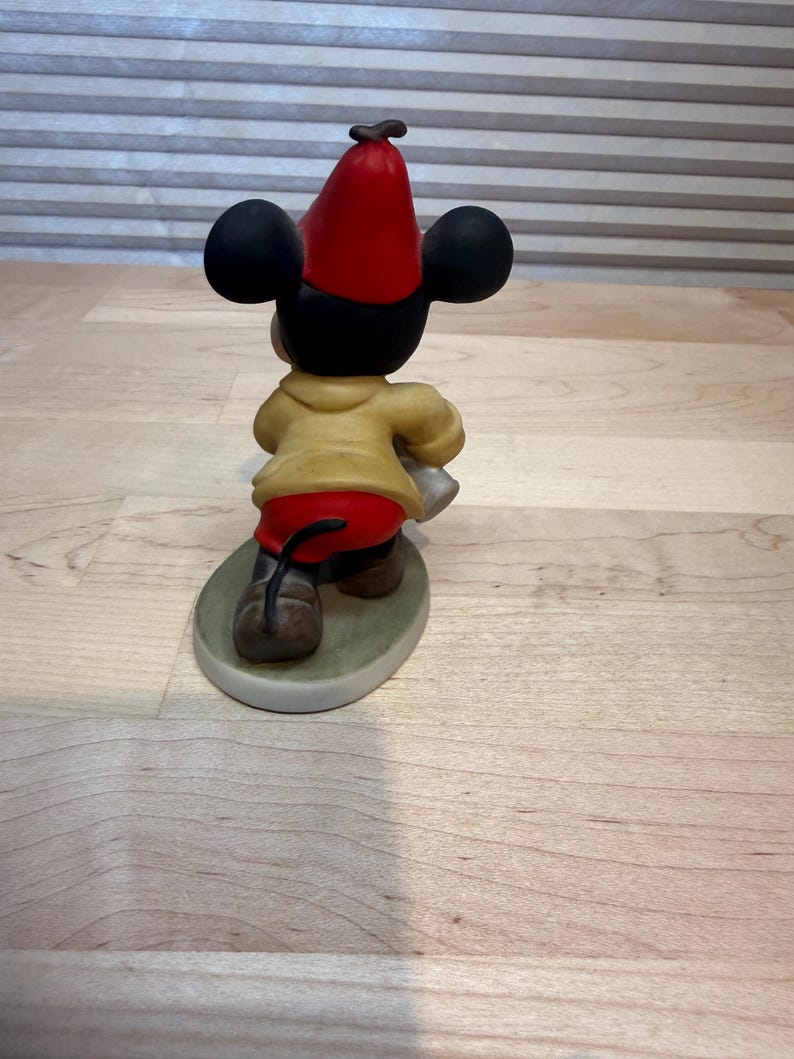 May include: A ceramic figurine of Mickey Mouse from the back, wearing a red hat, yellow jacket, and red shorts. The figure is standing on a green oval base. The figurine has black ears and brown shoes.