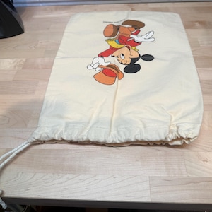 May include: A cream-colored drawstring bag featuring a colorful illustration of Mickey Mouse in a cowboy outfit. The cartoon character is depicted with a red shirt, yellow vest, brown pants, and a cowboy hat. The bag is made of a soft, light-colored fabric.