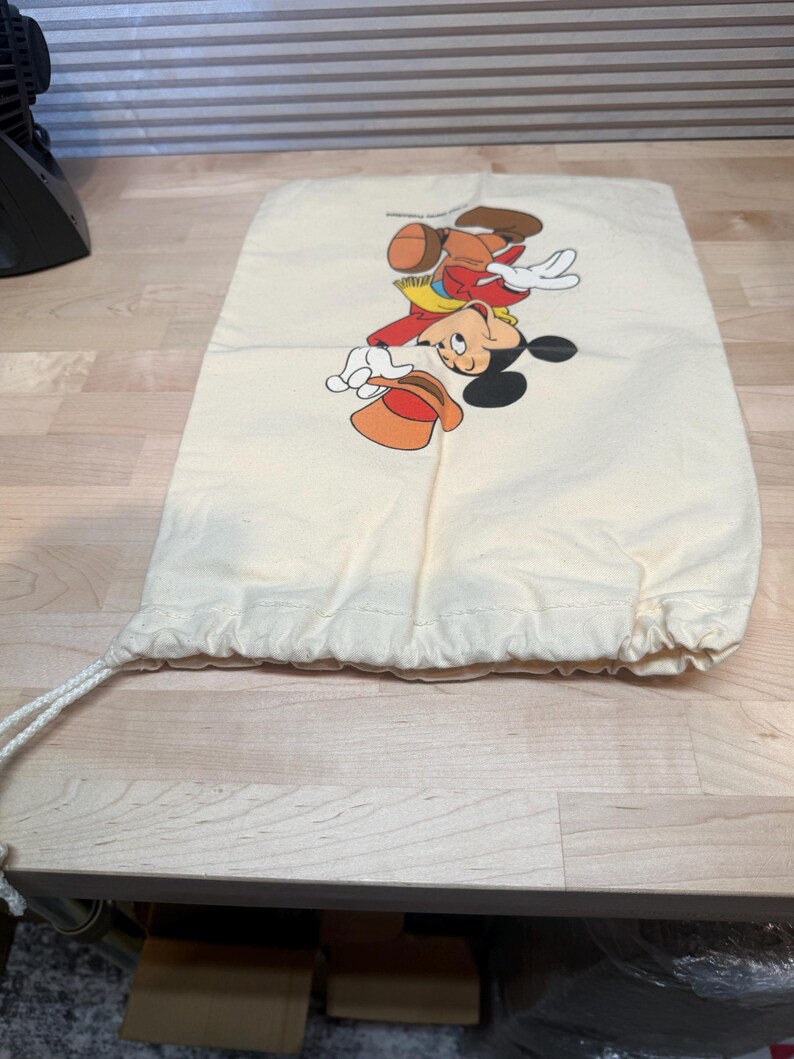 May include: A cream-colored drawstring bag featuring a colorful illustration of Mickey Mouse in a cowboy outfit. The cartoon character is depicted with a red shirt, yellow vest, brown pants, and a cowboy hat. The bag is made of a soft, light-colored fabric.