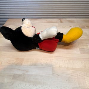 May include: A plush Mickey Mouse toy in a reclining pose. The toy has a black body, white gloves, a cream-coloured face, red shorts, and yellow shoes. The toy is lying on a light-coloured wooden surface.