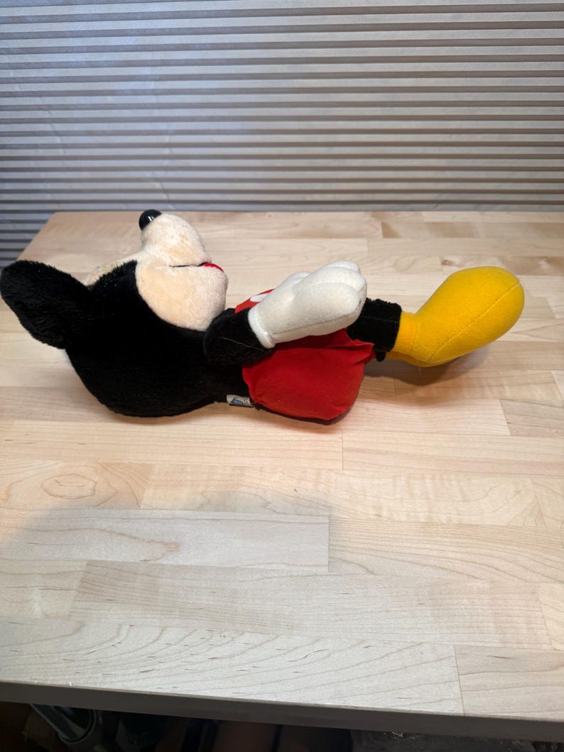 May include: A plush Mickey Mouse toy in a reclining pose. The toy has a black body, white gloves, a cream-coloured face, red shorts, and yellow shoes. The toy is lying on a light-coloured wooden surface.