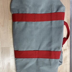 Puede incluir: Una bolsa de lona gris con correas rojas. La bolsa tiene un asa superior y dos correas laterales.