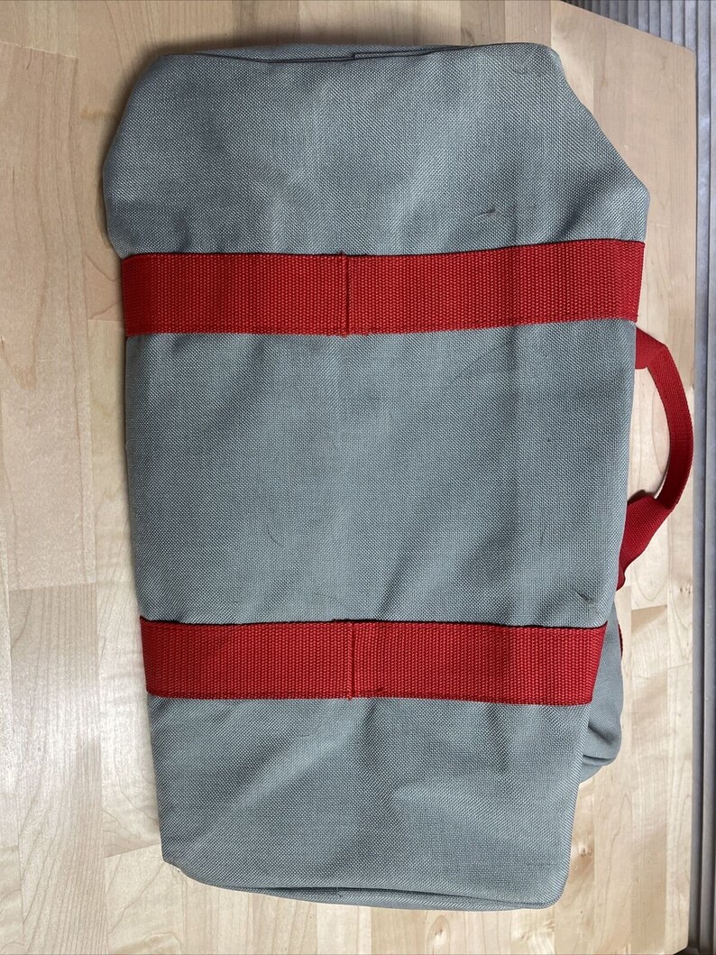 Puede incluir: Una bolsa de lona gris con correas rojas. La bolsa tiene un asa superior y dos correas laterales.
