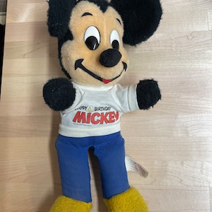 May include: A plush Mickey Mouse toy wearing a white t-shirt with the text "Happy Birthday Mickey" and blue trousers. The toy has black ears, a black nose, and yellow shoes.