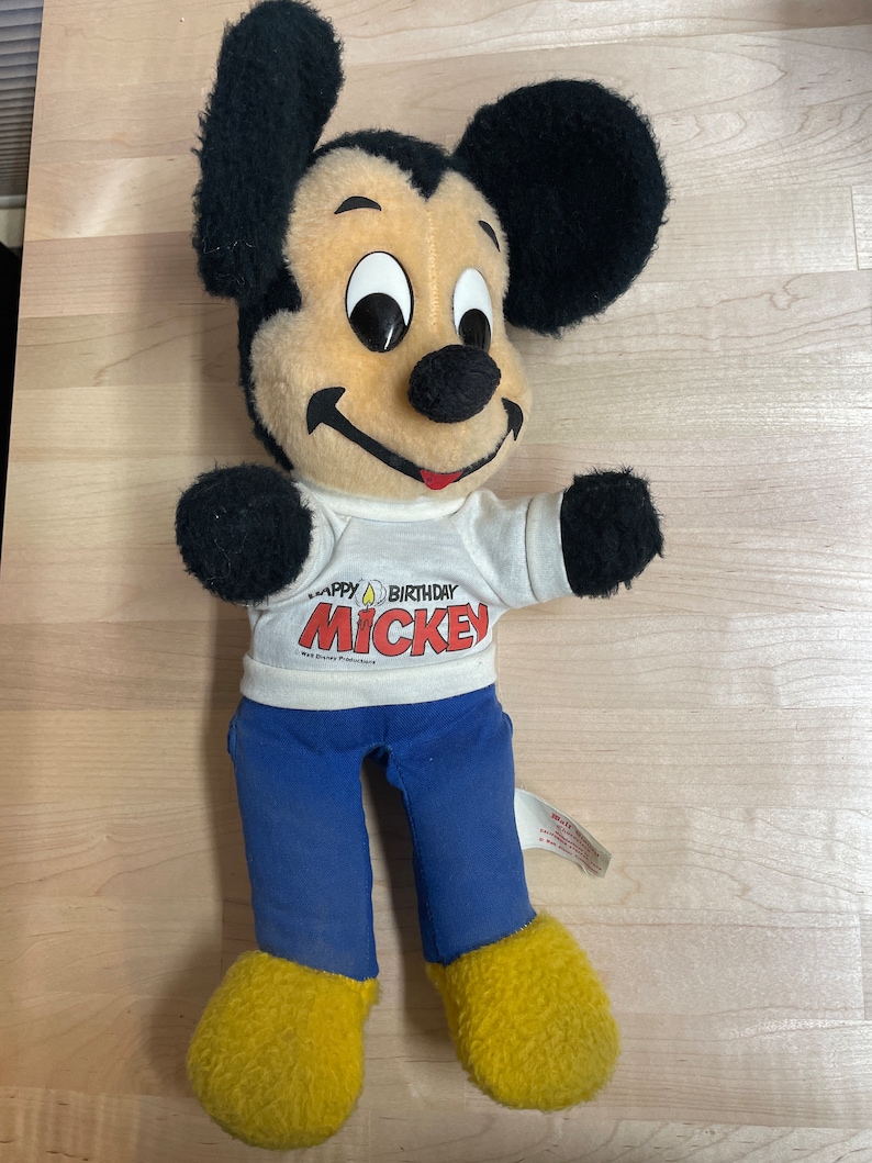 May include: A plush Mickey Mouse toy wearing a white t-shirt with the text "Happy Birthday Mickey" and blue trousers. The toy has black ears, a black nose, and yellow shoes.