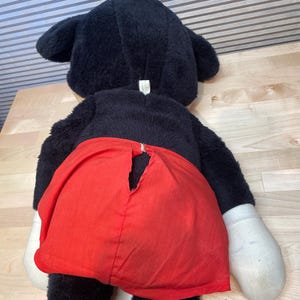 K&ouml;nnte beinhalten: Ein gro&szlig;es, vintage Mickey Mouse Pl&uuml;schtier, von hinten betrachtet. Das Pl&uuml;schtier hat schwarzes Fell, gro&szlig;e Ohren, wei&szlig;e Handschuhe und rote Shorts. Die Shorts haben einen Riss im Stoff. Das Spielzeug liegt auf einer hellen Holzoberfl&auml;che.