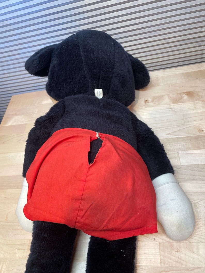 K&ouml;nnte beinhalten: Ein gro&szlig;es, vintage Mickey Mouse Pl&uuml;schtier, von hinten betrachtet. Das Pl&uuml;schtier hat schwarzes Fell, gro&szlig;e Ohren, wei&szlig;e Handschuhe und rote Shorts. Die Shorts haben einen Riss im Stoff. Das Spielzeug liegt auf einer hellen Holzoberfl&auml;che.
