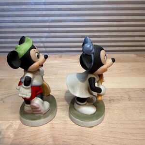 Puede incluir: Dos figuras de cer&aacute;mica de Mickey y Minnie Mouse con atuendo de tenis. Mickey lleva gorra verde, camisa blanca, pantalones cortos rojos y zapatos blancos, sosteniendo una raqueta. Minnie viste vestido blanco y lazo azul, sosteniendo una raqueta.