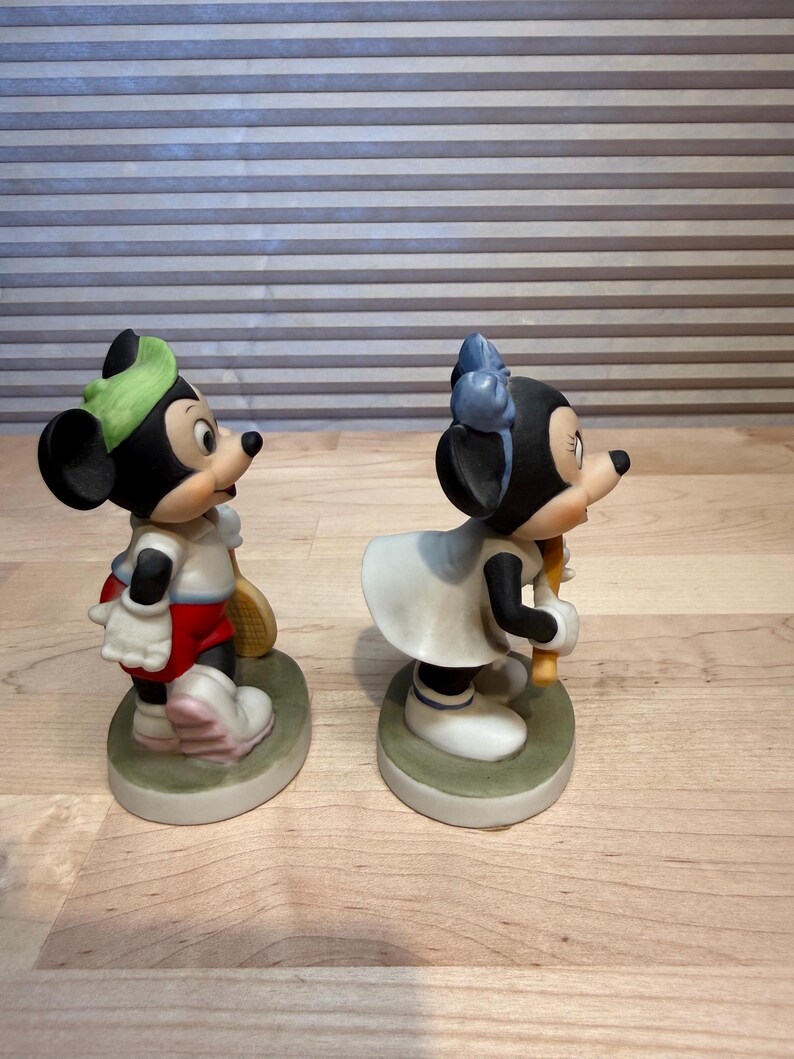 Puede incluir: Dos figuras de cer&aacute;mica de Mickey y Minnie Mouse con atuendo de tenis. Mickey lleva gorra verde, camisa blanca, pantalones cortos rojos y zapatos blancos, sosteniendo una raqueta. Minnie viste vestido blanco y lazo azul, sosteniendo una raqueta.