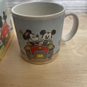 K&ouml;nnte beinhalten: Keramikbecher mit Mickey und Minnie Mouse in einem roten Auto. Der Becher hat einen hellblauen Hintergrund mit einem cremefarbenen Rand und Henkel. Das Design umfasst eine gelbe Blume und einen beigefarbenen Sockel.
