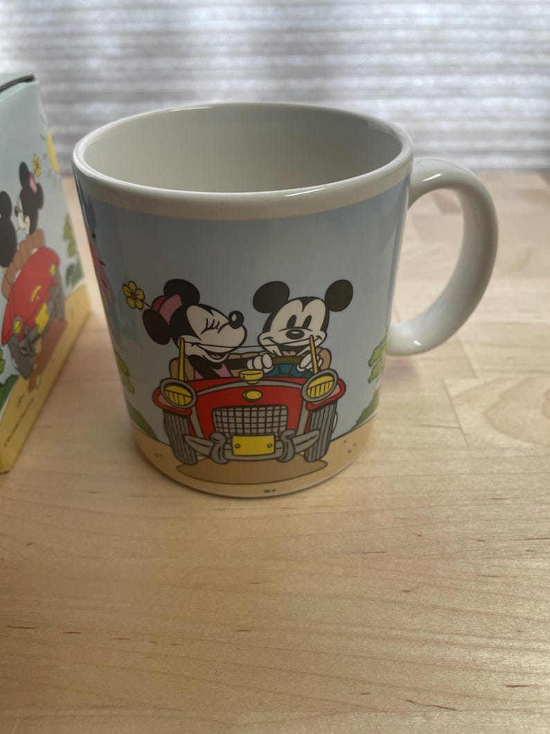 K&ouml;nnte beinhalten: Keramikbecher mit Mickey und Minnie Mouse in einem roten Auto. Der Becher hat einen hellblauen Hintergrund mit einem cremefarbenen Rand und Henkel. Das Design umfasst eine gelbe Blume und einen beigefarbenen Sockel.