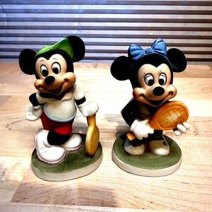 Puede incluir: Dos figuras de cer&aacute;mica de Mickey y Minnie Mouse, cada uno sosteniendo una raqueta de tenis. Mickey lleva un gorro verde, camisa blanca, pantalones cortos rojos y zapatos blancos. Minnie lleva un lazo azul y un vestido negro. Ambos est&aacute;n sobre bases verdes.
