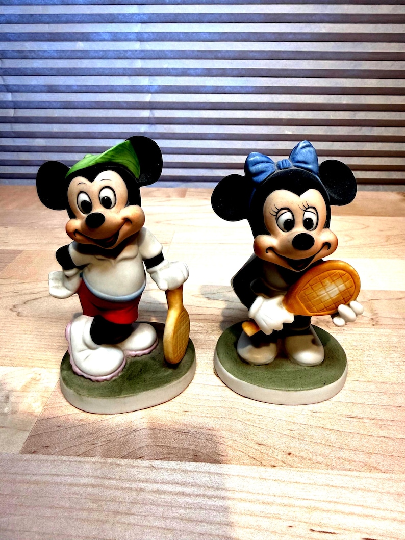 Puede incluir: Dos figuras de cer&aacute;mica de Mickey y Minnie Mouse, cada uno sosteniendo una raqueta de tenis. Mickey lleva un gorro verde, camisa blanca, pantalones cortos rojos y zapatos blancos. Minnie lleva un lazo azul y un vestido negro. Ambos est&aacute;n sobre bases verdes.