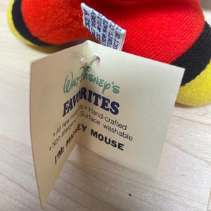 Puede incluir: Un peluche de Mickey Mouse con cuerpo rojo, detalles negros y pies amarillos. La etiqueta adjunta dice "Walt Disney's Favorites" y "I'M: MICKEY MOUSE."