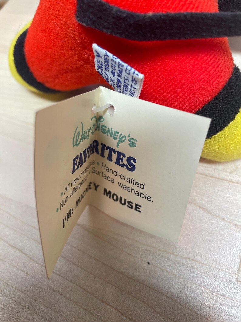 Puede incluir: Un peluche de Mickey Mouse con cuerpo rojo, detalles negros y pies amarillos. La etiqueta adjunta dice "Walt Disney's Favorites" y "I'M: MICKEY MOUSE."