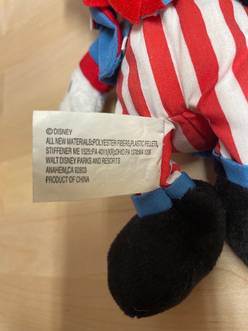 K&ouml;nnte beinhalten: Ein rotes, wei&szlig;es und blaues Pl&uuml;schtier mit schwarzem Gesicht und schwarzen Schuhen. Das Spielzeug hat ein Etikett mit der Aufschrift "DISNEY ALL NEW MATERIALS:POLYESTER FIBERS, PLASTIC PELLETS, STIFFENER ME 1525:PA 4011 (KR) OHIO PA 1378:MA 1336 WALT DISNEY PARKS AND RESORTS ANAHEIM,CA 92803 PRODUCT OF CHINA"