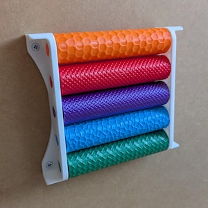 Könnte beinhalten: Ein weißer Wandhalter aus Kunststoff mit fünf zylindrischen Rollen in Orange, Rot, Lila, Blau und Grün. Jede Rolle weist ein eigenes strukturiertes Oberflächenmuster auf. Der weiße Rahmen ist mit zwei sichtbaren Schrauben an der linken Seite befestigt.