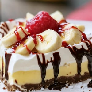 Puede incluir: Un postre con capas de chocolate, crema de vainilla y una base de galletas graham, cubierto con rodajas de plátano, una frambuesa y salsa de chocolate.