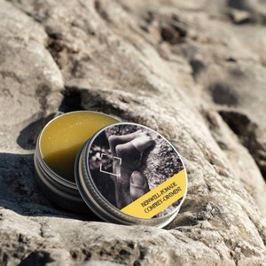 Puede incluir: Una lata de plata con un ungüento amarillo en su interior. La tapa de la lata tiene una foto en blanco y negro de una mano sobre una roca y el texto "BEINWELL POMADE COMFREY OINTMENT".