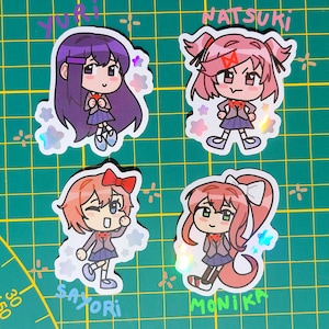 Puede incluir: Cuatro pegatinas holográficas con personajes de estilo anime. Cada pegatina representa a una chica diferente con peinados y atuendos únicos. Los nombres "Yuri", "Natsuki", "Sayori" y "Monika" están escritos encima o debajo de cada personaje.
