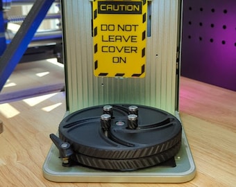 May include: A silver and green 3D printer with a black rotating platform. The printer has a yellow warning sign that reads "CAUTION DO NOT LEAVE COVER ON".