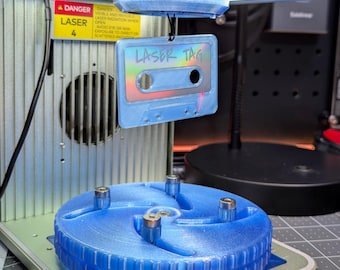 May include: A blue cassette tape with the text "LASER TAG" is suspended in front of a laser engraving machine. The machine has a green top and a blue base. A warning label is visible. The cassette tape has a holographic effect.