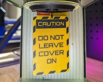May include: A yellow caution sign with black text that reads "CAUTION DO NOT LEAVE COVER ON". The sign is hanging from a black and silver device.