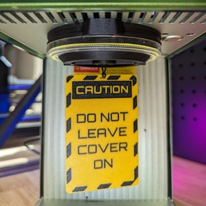 Op de afbeelding: Een geel waarschuwingsbord met zwarte tekst die "CAUTION DO NOT LEAVE COVER ON" luidt. Het bord hangt aan een zwart en zilver apparaat.