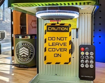 May include: A green laser engraver with a yellow caution sign that reads "Do Not Leave Cover On". The engraver has a circular platform and a remote control labeled "DIM CONTROLLER".