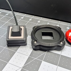 Op de afbeelding: Drie 3D-geprinte objecten: een zwarte vierkante knop met een grijze knop, een zwarte ronde basis met een vierkante uitsparing en een rode ronde knop met het woord "FIRE" in wit.