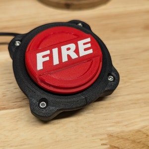 Op de afbeelding: Een rode knop met het woord "FIRE" erop gedrukt in wit. De knop is bevestigd aan een zwarte kunststof basis met een snoer.