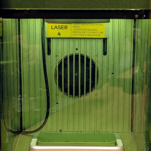 May include: Close-up of a green laser cutter with a warning label that reads "LASER RADIATION WHEN OPEN AVOID EYE OR SKIN EXPOSURE TO DIRECT OR SCATTERED RADIATION". The label is yellow with black text. The laser cutter has a small fan in the back.