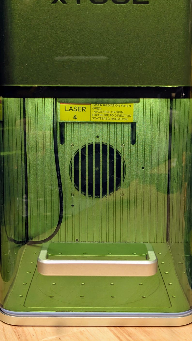 May include: Close-up of a green laser cutter with a warning label that reads "LASER RADIATION WHEN OPEN AVOID EYE OR SKIN EXPOSURE TO DIRECT OR SCATTERED RADIATION". The label is yellow with black text. The laser cutter has a small fan in the back.