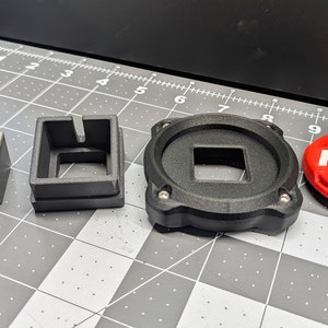 Op de afbeelding: Vier 3D-geprinte objecten: een zwarte vierkant met een vierkant gat, een zwart rond object met een vierkant gat, een zwarte vierkant met een toetsdop en de letter "X" erop en een rode ronde knop met het woord "FIRE" erop.