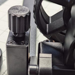 K&ouml;nnte beinhalten: Nahaufnahme eines schwarzen 3D-gedruckten Objekts mit einem zahnradf&ouml;rmigen Rad und einem Knopf. Das Objekt ist auf einem schwarzen Sockel mit vier Schrauben montiert.