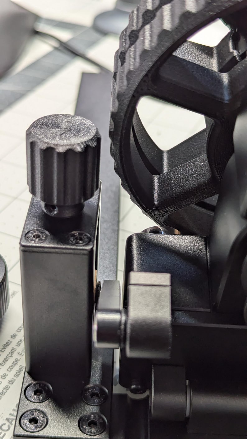 K&ouml;nnte beinhalten: Nahaufnahme eines schwarzen 3D-gedruckten Objekts mit einem zahnradf&ouml;rmigen Rad und einem Knopf. Das Objekt ist auf einem schwarzen Sockel mit vier Schrauben montiert.