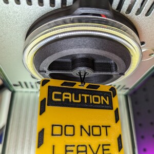 Op de afbeelding: Een geel en zwart waarschuwingsbord met de tekst "CAUTION DO NOT LEAVE" hangend aan een zwart touwtje. Het bord hangt aan een machine met een zwarte en zilveren ronde basis.