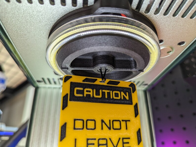 Op de afbeelding: Een geel en zwart waarschuwingsbord met de tekst "CAUTION DO NOT LEAVE" hangend aan een zwart touwtje. Het bord hangt aan een machine met een zwarte en zilveren ronde basis.