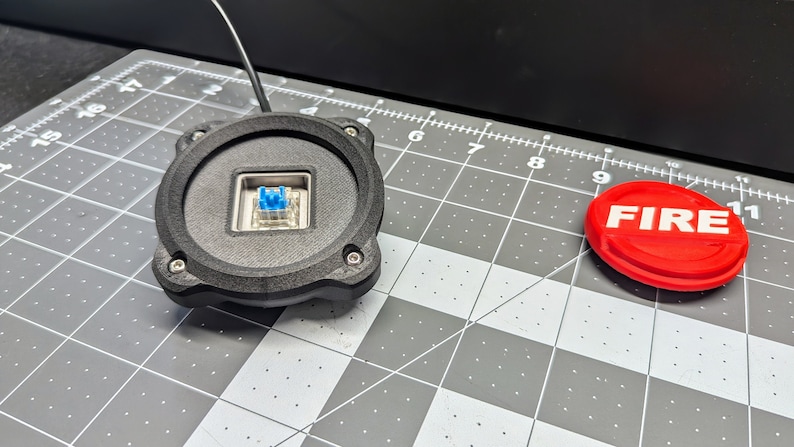 Op de afbeelding: Een zwart rond apparaat met een blauwe mechanische toetsenbordschakelaar in het midden. Een ronde rode knop met het woord "FIRE" in witte letters bevindt zich rechts.