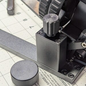 K&ouml;nnte beinhalten: Nahaufnahme einer schwarzen Metallmaschine mit zwei Zahnr&auml;dern und einem Knopf. Die Maschine steht auf einer wei&szlig;en, karierten Oberfl&auml;che, auf der die Zahlen 21 und 22 sichtbar sind. Der Text auf der Maschine lautet: "A CAUTION Avoid cutting with excessive pressure to avoid damaging the mat. Heat and direct sunlight. A MISE EN GARDE &Eacute;vitez de reprendre au m&ecirc;me endroit ou en exer&ccedil;ant une pression pour ne pas fendre le tapis de coupe. &Eacute;vitez le chaleur et la lumi&egrave;re directe du soleil. ▲ PRECAUCI mismo lugar o Evite el fo"