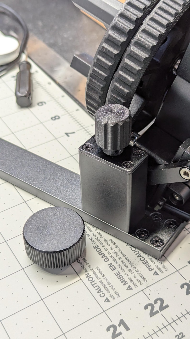 K&ouml;nnte beinhalten: Nahaufnahme einer schwarzen Metallmaschine mit zwei Zahnr&auml;dern und einem Knopf. Die Maschine steht auf einer wei&szlig;en, karierten Oberfl&auml;che, auf der die Zahlen 21 und 22 sichtbar sind. Der Text auf der Maschine lautet: "A CAUTION Avoid cutting with excessive pressure to avoid damaging the mat. Heat and direct sunlight. A MISE EN GARDE &Eacute;vitez de reprendre au m&ecirc;me endroit ou en exer&ccedil;ant une pression pour ne pas fendre le tapis de coupe. &Eacute;vitez le chaleur et la lumi&egrave;re directe du soleil. ▲ PRECAUCI mismo lugar o Evite el fo"
