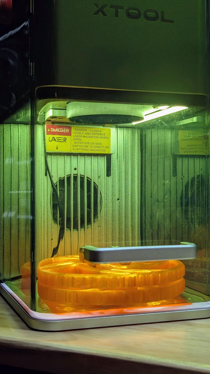 May include: A close-up of a 3D printed object inside a clear enclosure. The object is orange and appears to be a gear or a wheel. A warning label is visible on the enclosure, stating "DANGER - CLASS 4 VISIBLE AND INVISIBLE LASER RADIATION WHEN OPEN AVOID EYE OR SKIN EXPOSURE TO DIRECT OR SCATTERED RADIATION".