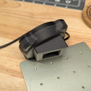 Op de afbeelding: Een zwart 3D-geprint object met een rechthoekige basis en een ronde bovenkant. Het object is verbonden met een zwarte kabel en ligt op een grijs metalen oppervlak.