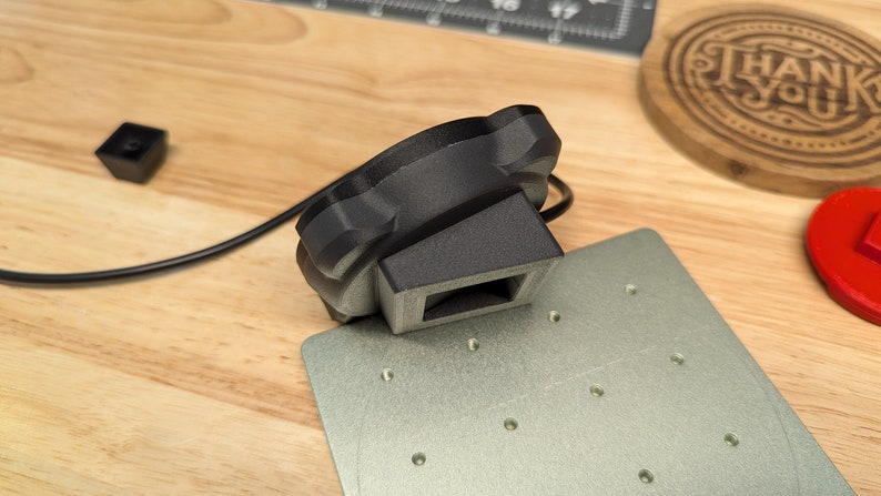 Op de afbeelding: Een zwart 3D-geprint object met een rechthoekige basis en een ronde bovenkant. Het object is verbonden met een zwarte kabel en ligt op een grijs metalen oppervlak.