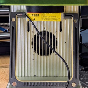 May include: Close-up of a laser engraver with a warning label that reads "LASER RADIATION WHEN OPEN AVOID EYE OR SKIN EXPOSURE TO DIRECT OR SCATTERED RADIATION". The laser is labeled "LASER 4".