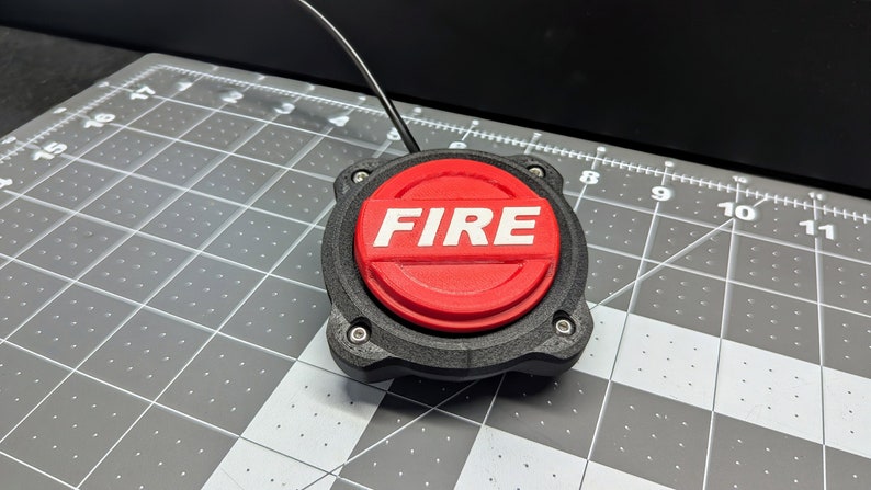 Op de afbeelding: Een rode drukknop met het woord "FIRE" in wit erop gedrukt. De knop is gemonteerd op een zwarte kunststof basis met vier schroeven.
